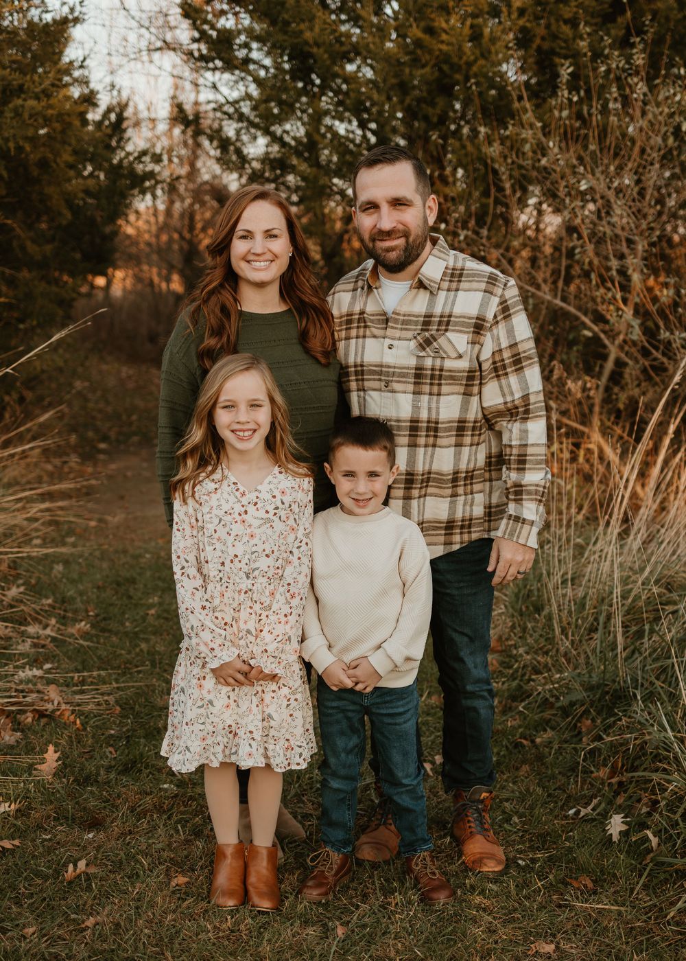 A family poses together in a rustic outdoor setting at sunset among tall grass and trees.