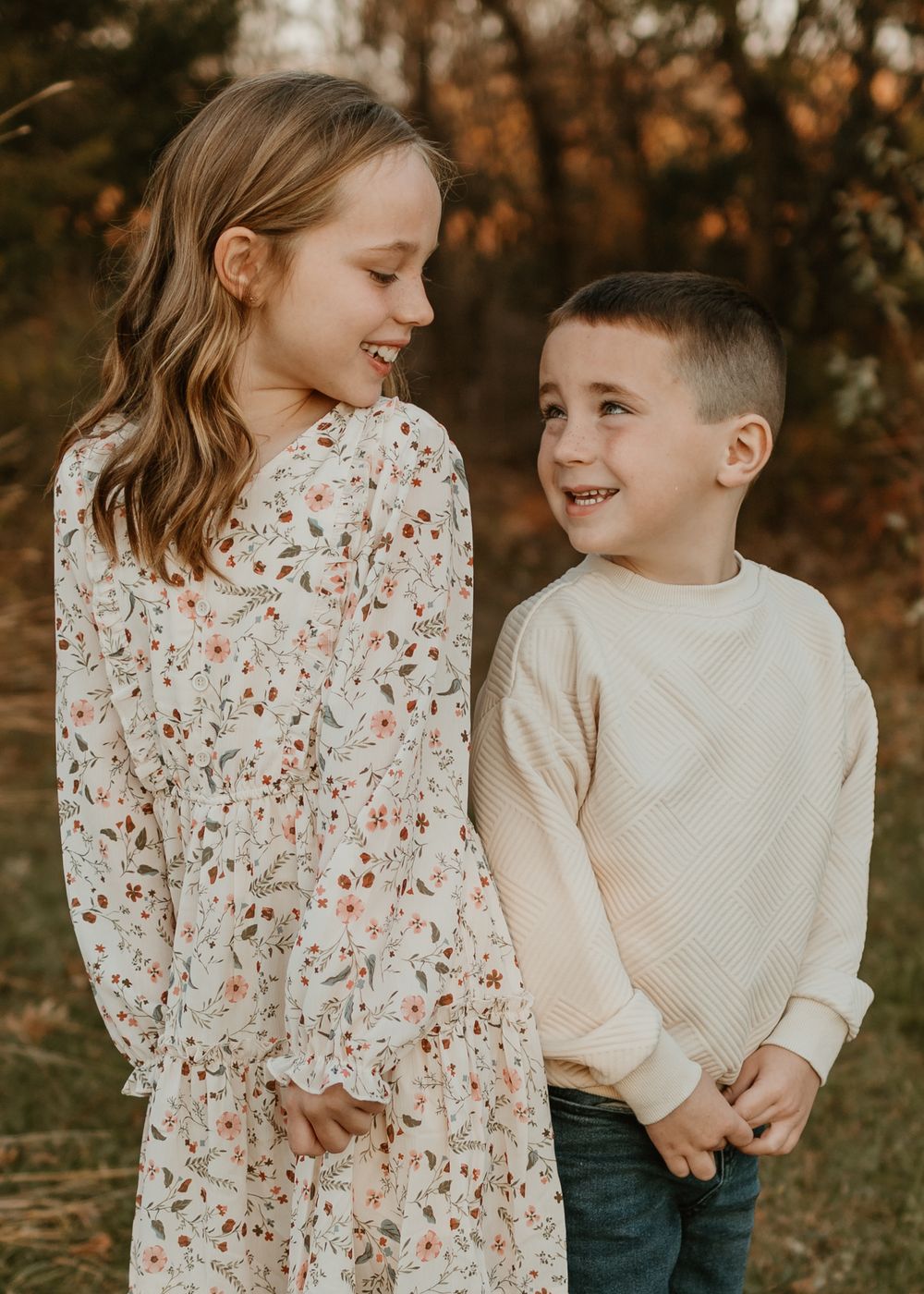 Two children share a playful moment together among autumn trees in warm golden light.