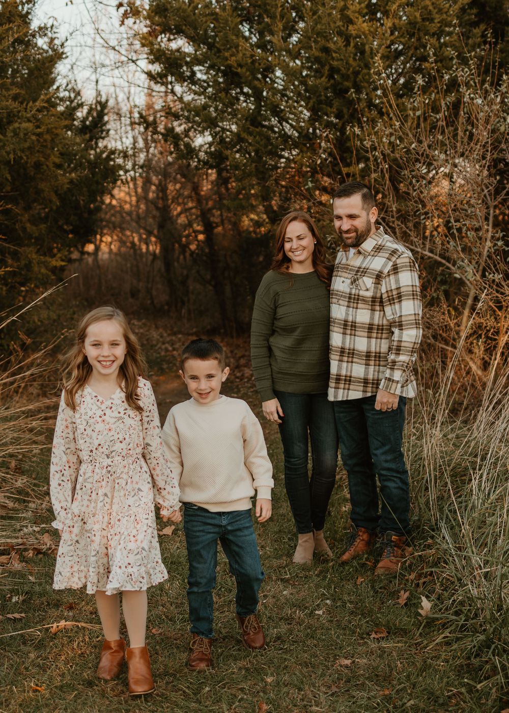 Family wearing coordinated fall outfits stands together in outdoor setting with autumn foliage at sunset.