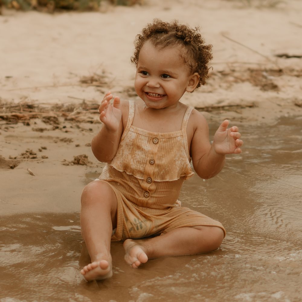 Joyful toddler in tan romper sits and plays on sandy beach in sepia-toned photograph.
