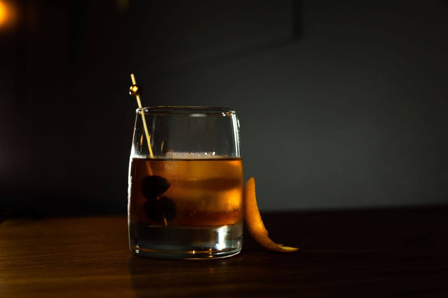 Elegant Old Fashioned cocktail with orange peel garnish on dark wooden bar surface.
