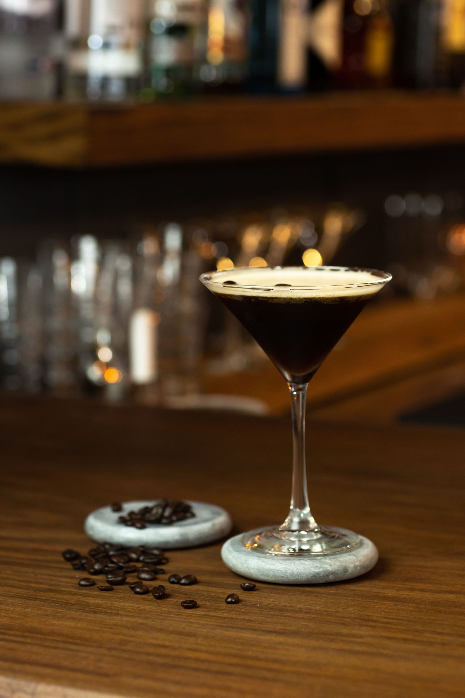 An elegant espresso martini cocktail in a martini glass garnished with coffee beans on a wooden bar counter.