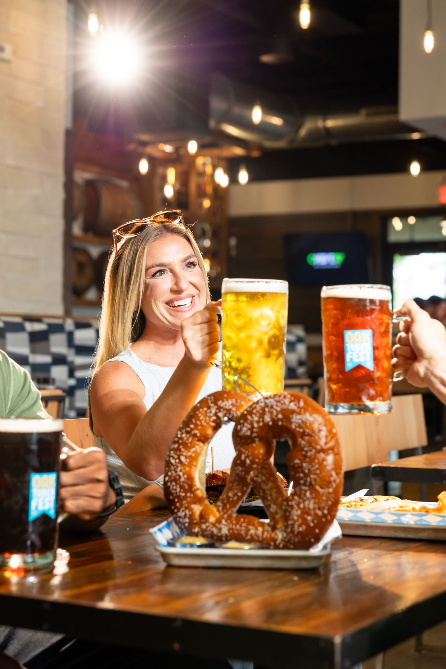 Smiling blonde woman toasting with beer mugs beside large pretzel at a brewery bar.