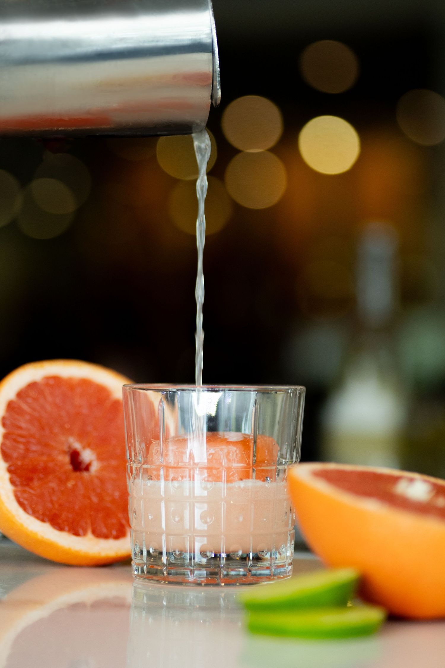 Bartender pouring cocktail shaker into glass with fresh grapefruit and lime garnish.