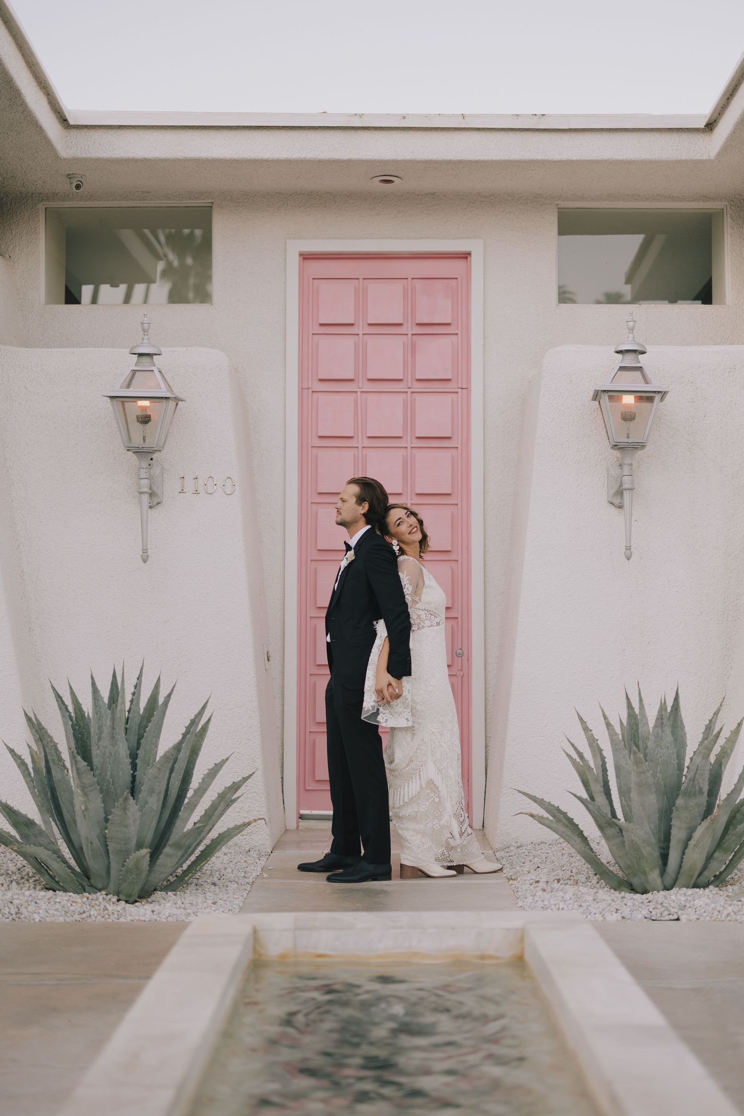 Wedding couple poses back-to-back in front of a vibrant pink door flanked by agave plants.