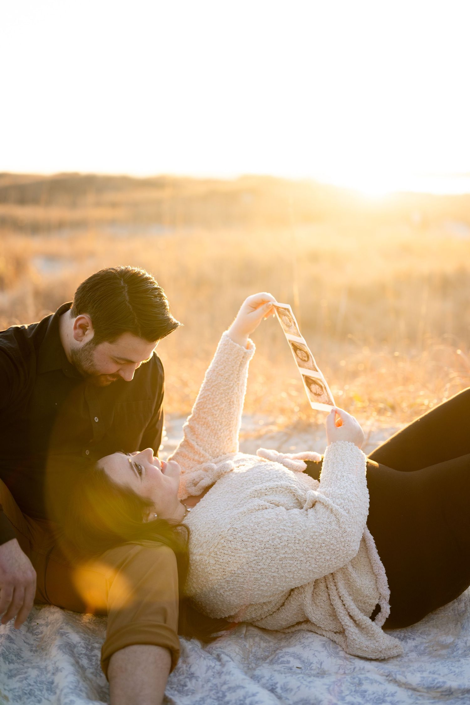 A couple lies together on a blanket in a golden sunlit field during sunset, creating a romantic silhouette.