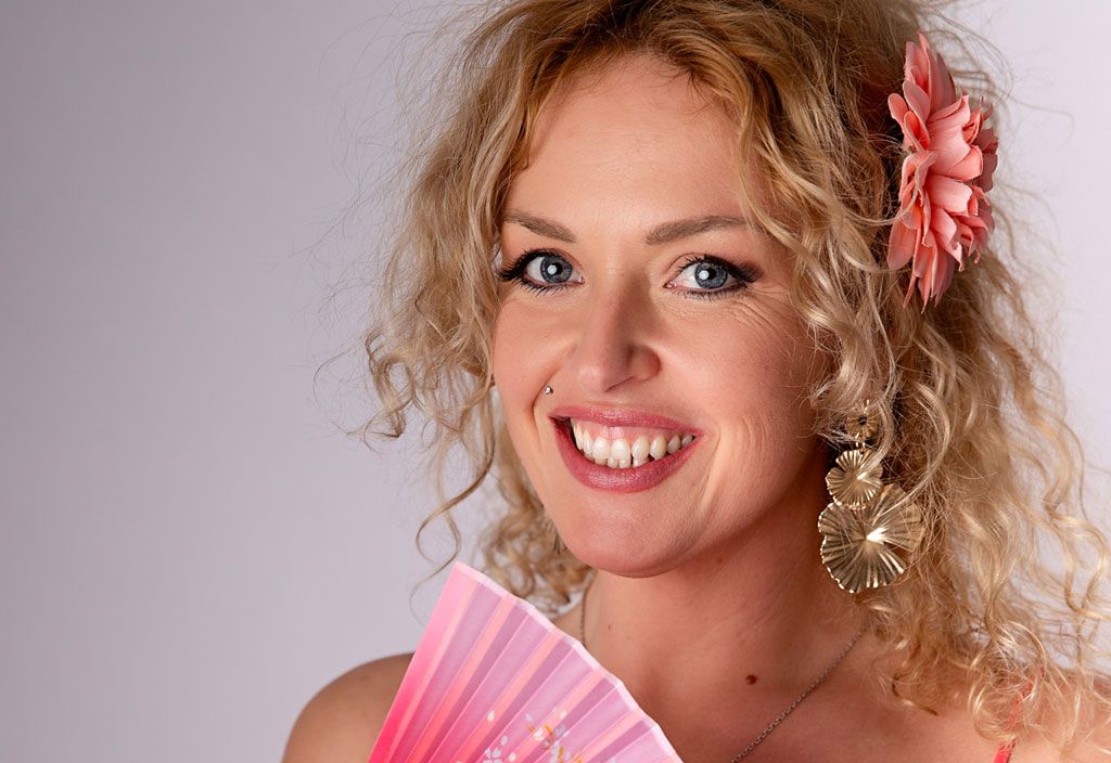 Woman in coral top holds decorative pink Asian folding fan while wearing a pink flower in her curly blonde hair.