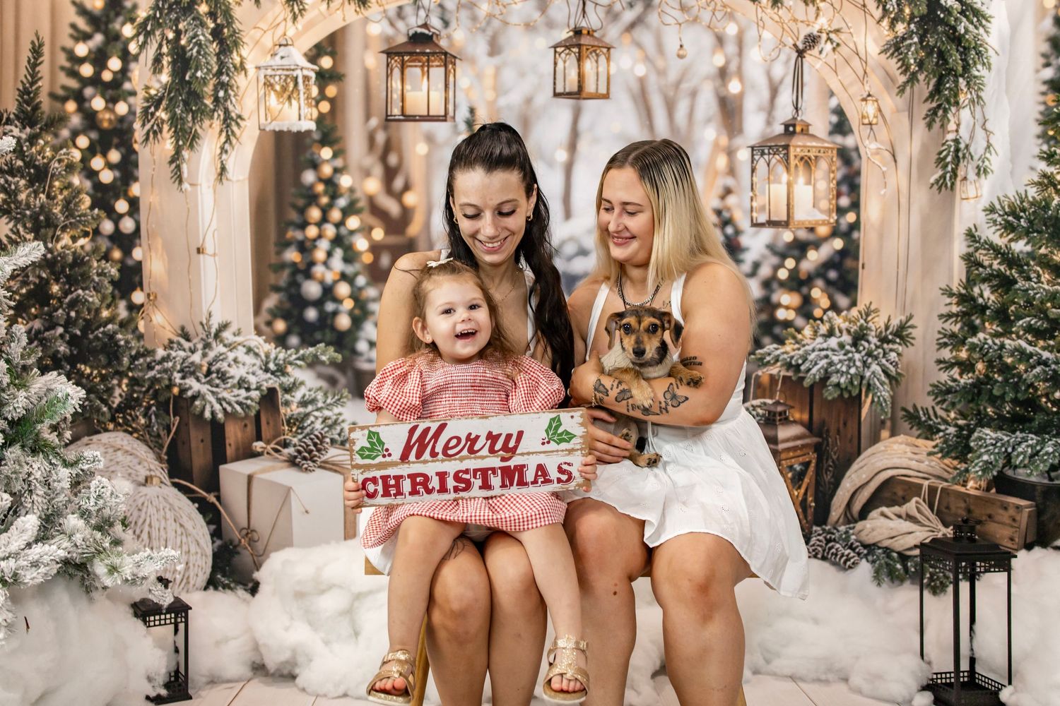Two women with toddler and dog holding Merry Christmas sign in holiday decor.