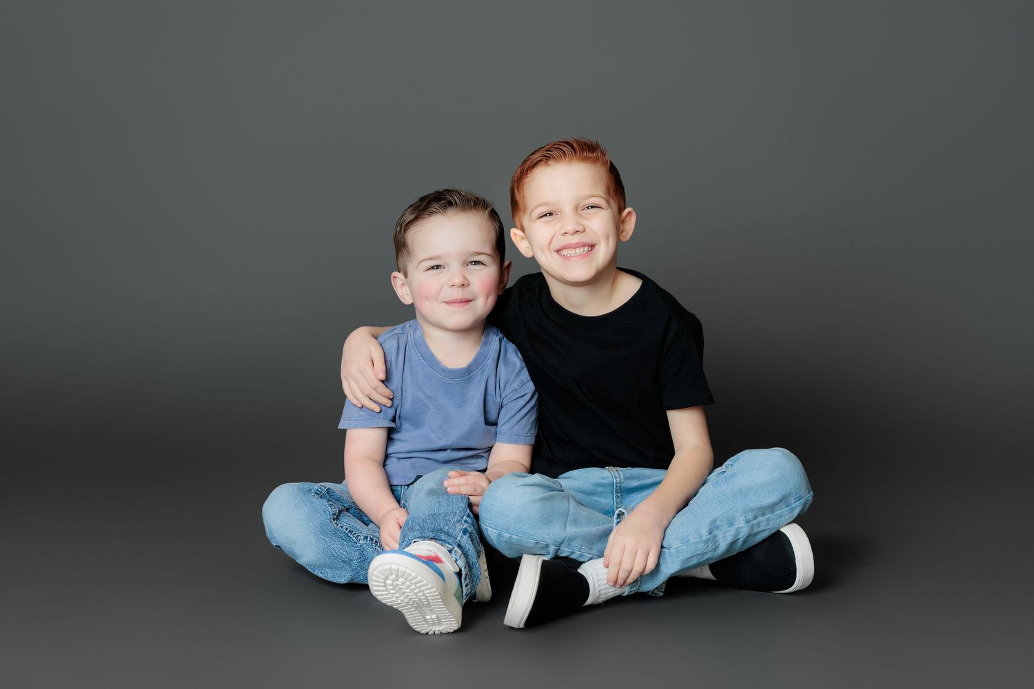 Two children sit together on a dark gray studio floor wearing casual clothing.
