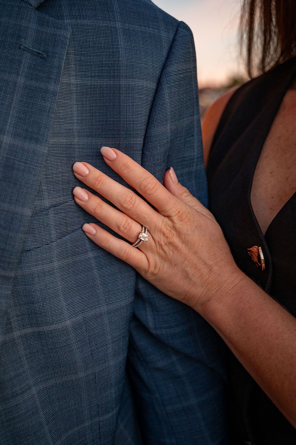 Close up of an engagement ring being shown on a hand against a blue suit jacket.