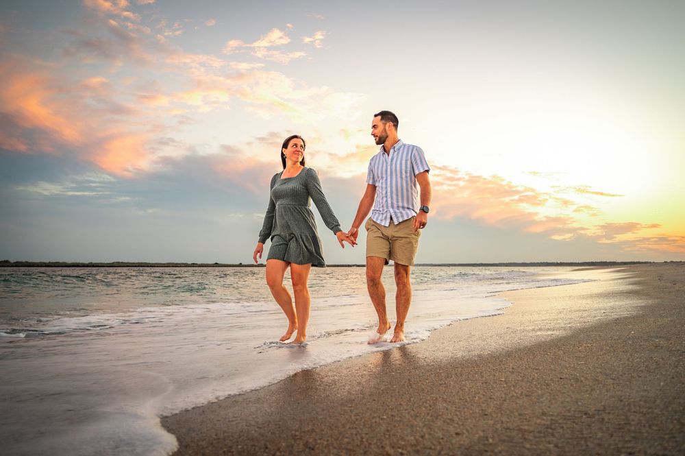 A couple walks hand in hand along a beach at sunset with pink clouds in the sky.