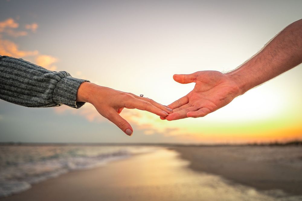 Two hands reaching toward each other against a golden sunset beach backdrop with ocean waves in the background.