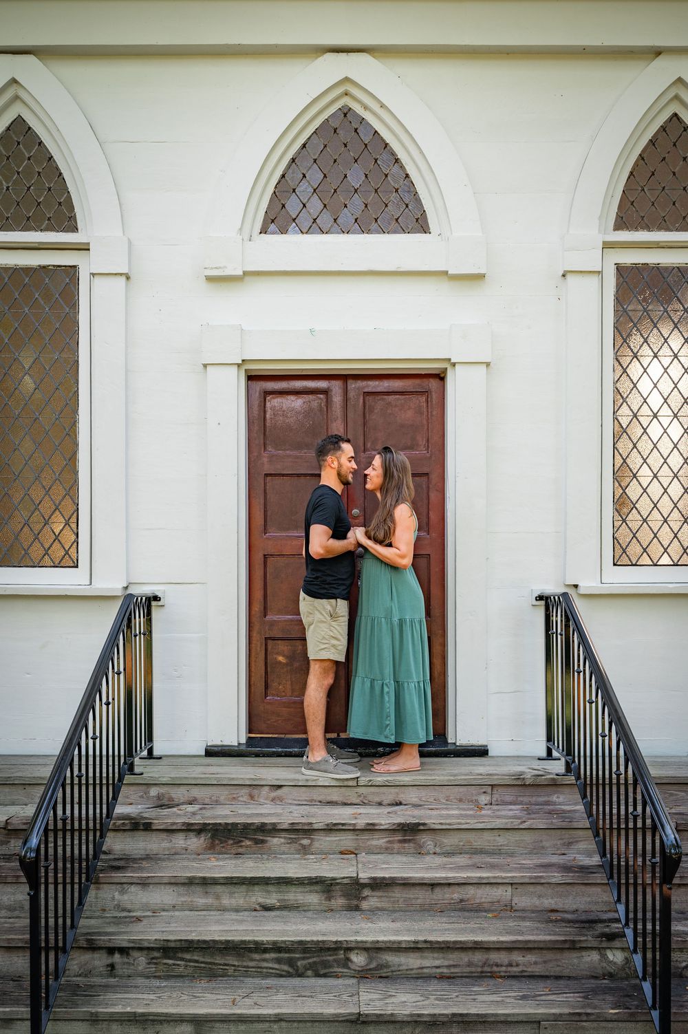 A couple shares a romantic moment on the steps of a historic church with Gothic arched windows and wooden doors.