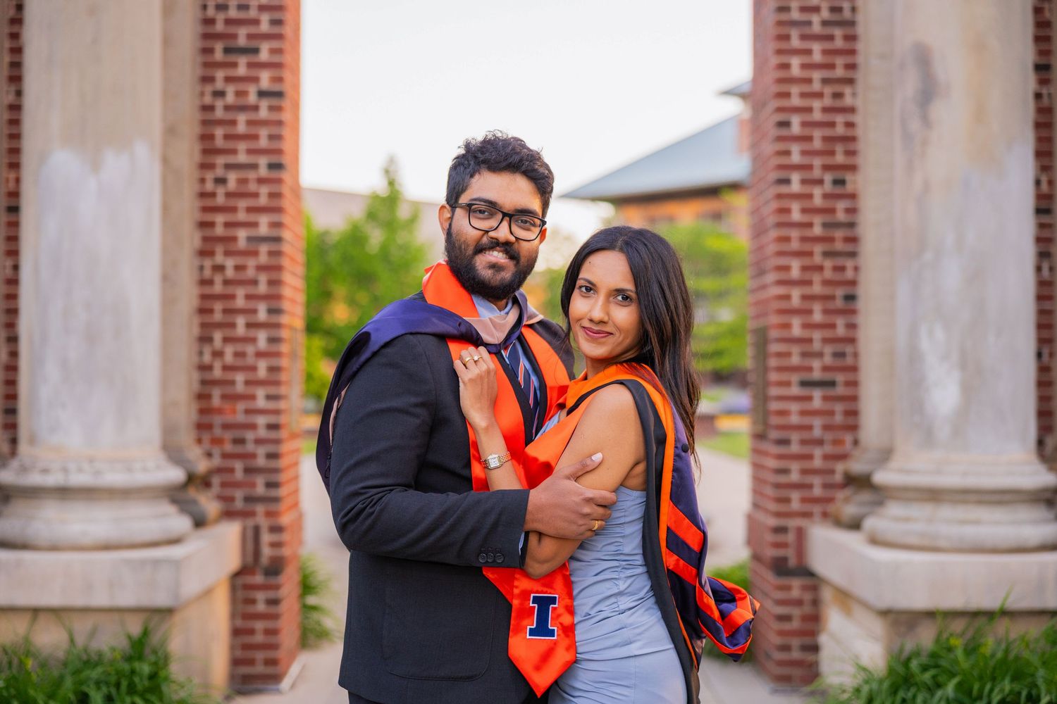 A UIUC graduating couple posed together in front of Hallene Gateway wearing graduation attire