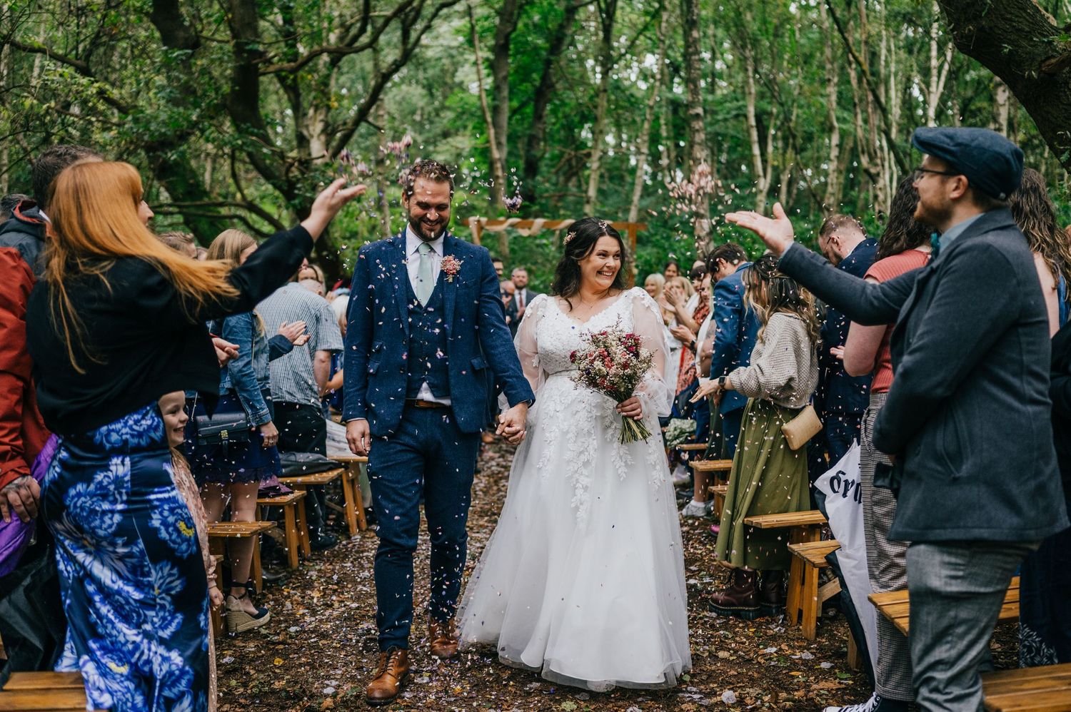 Bride and groom walk down outdoor woodland aisle as guests throw blue confetti at a forest wedding ceremony.