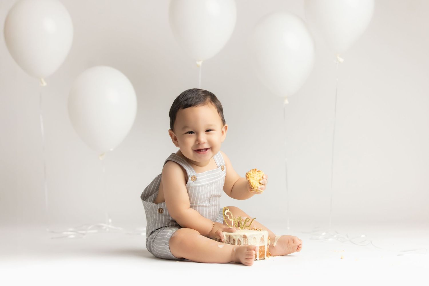 Smiling baby boy sitting on white floor holding a cupcake, surrounded by white balloons during first birthday cake smash.