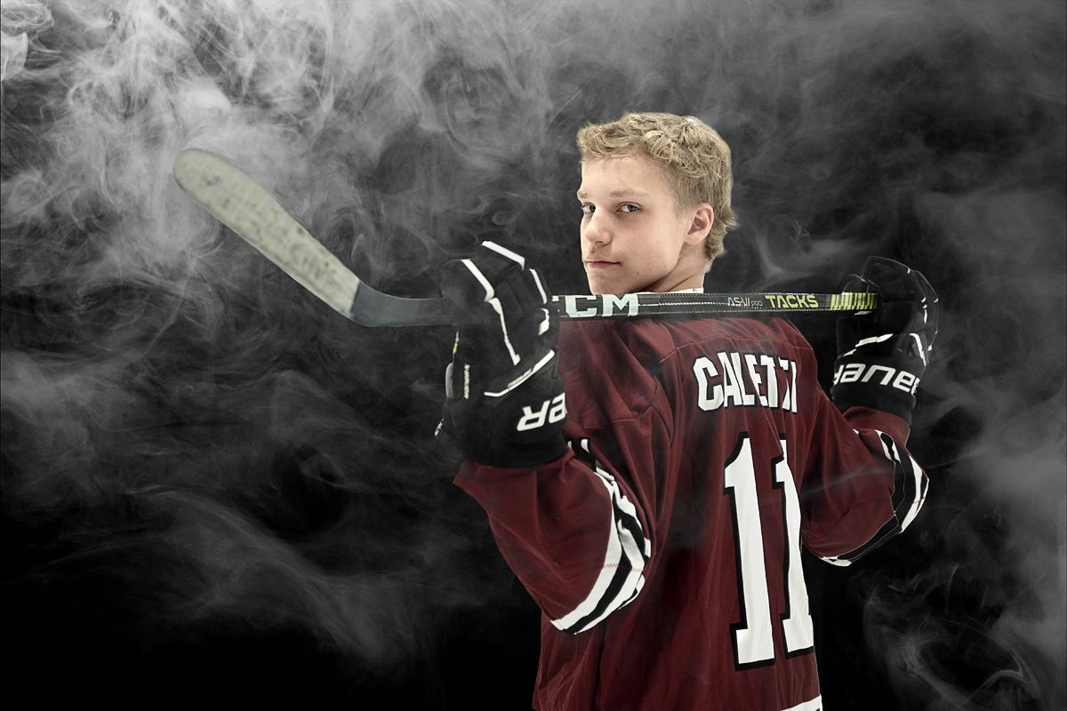 portrait of cool teen boy in hockey gear holding hockey stick over shoulders while looking at carmera with dark background and smoke effect*