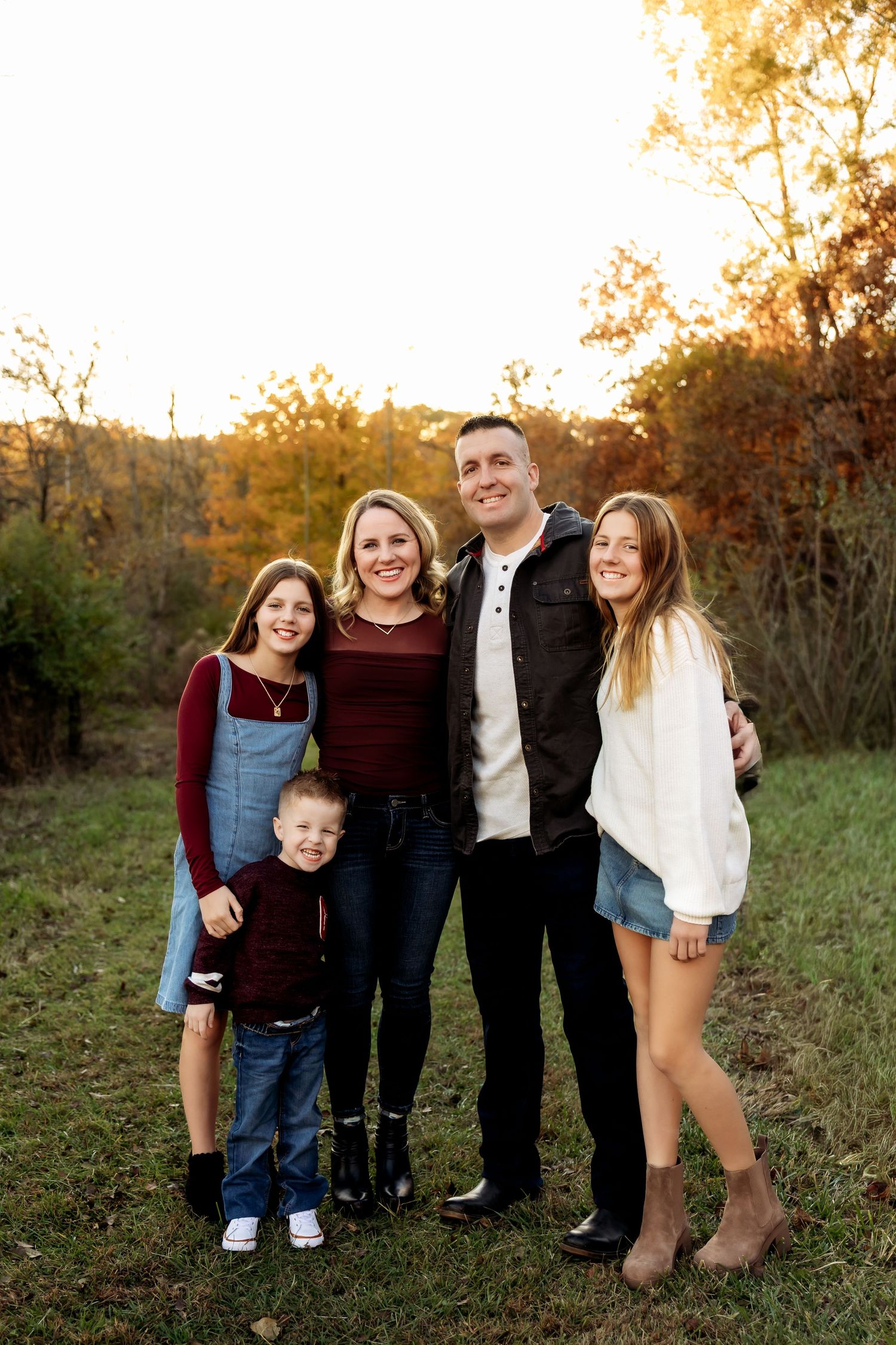 Family of five smiles together in golden sunset light during outdoor fall portrait session.