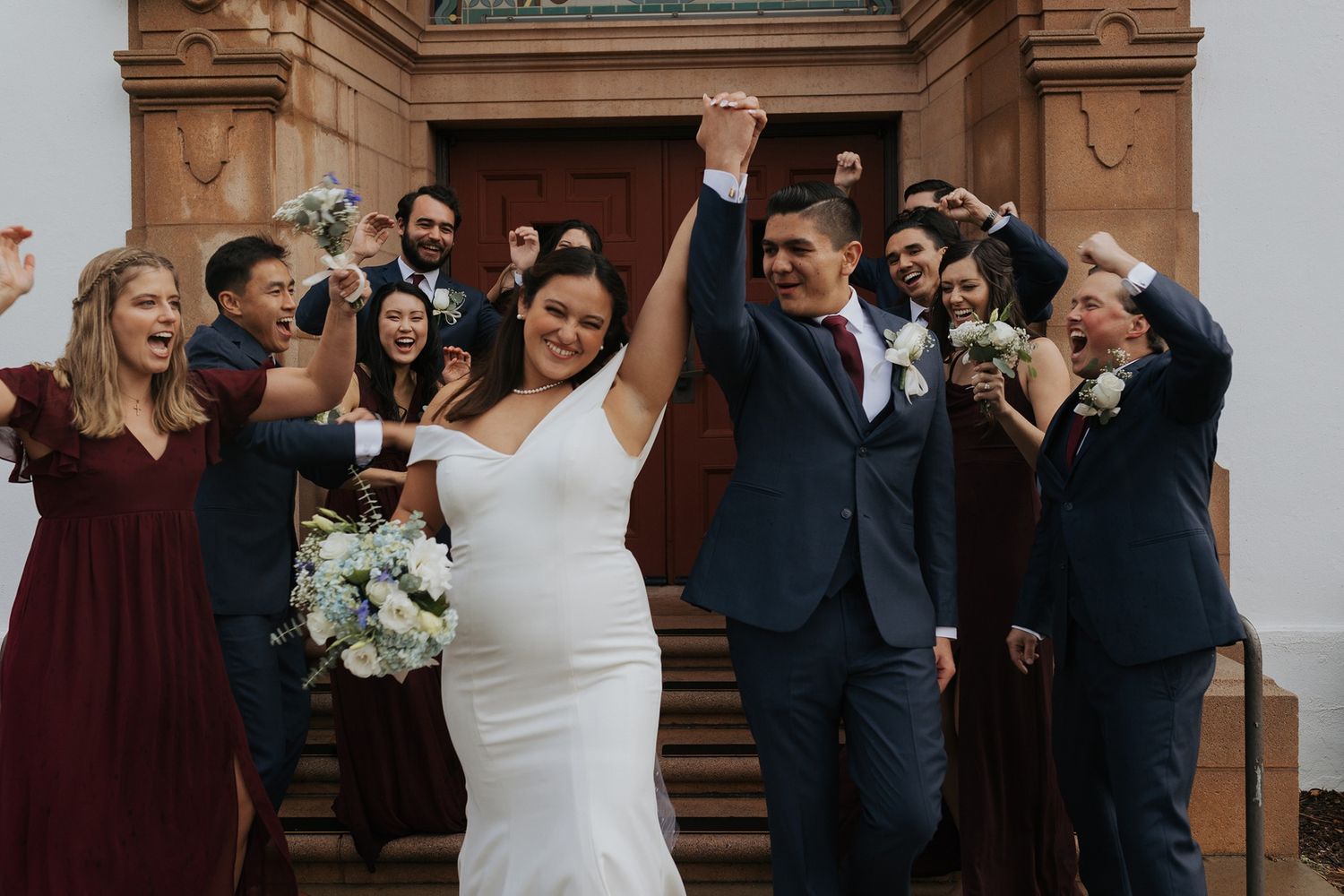 Wedding party celebrates joyfully outside historic stone building with ornate architecture.
