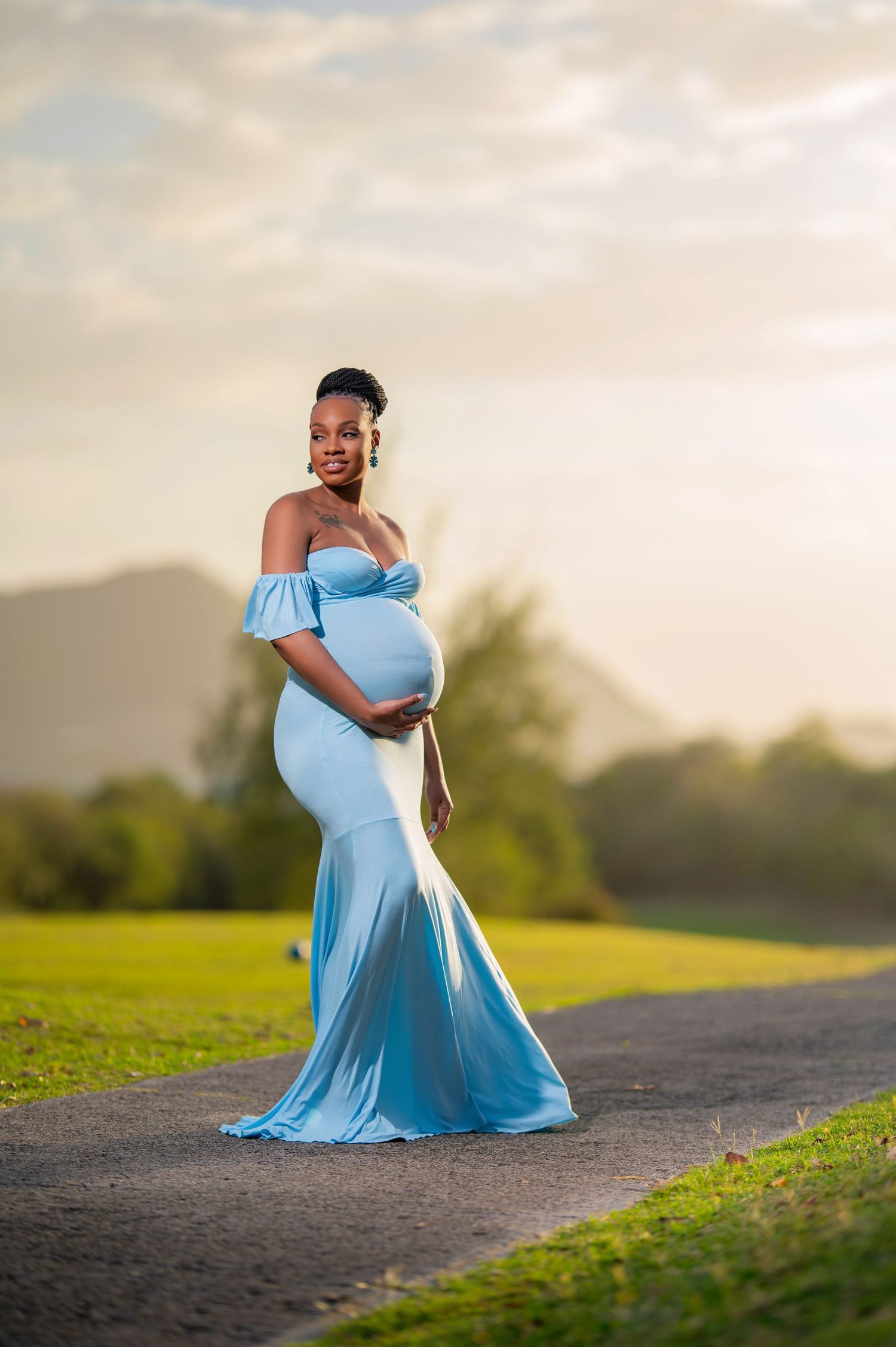 A woman in a flowing light blue dress poses gracefully on a grassy hillside during golden hour.