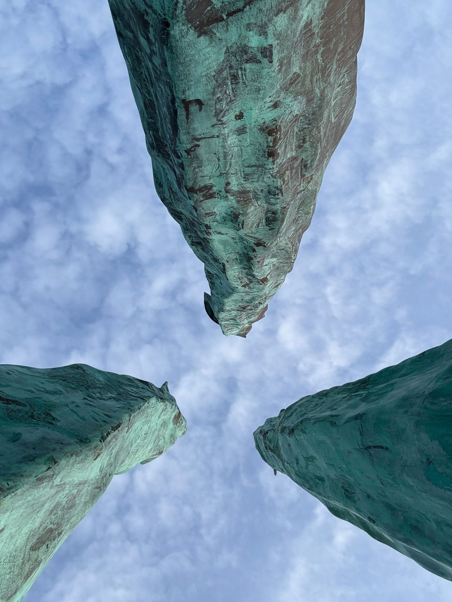 Looking up at three weathered green copper sculptures converging against a dramatic cloudy sky.