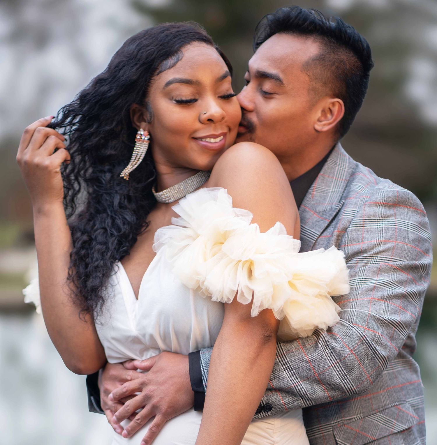 Romantic couple embraces in formal attire during outdoor photoshoot with soft lighting and blurred background.