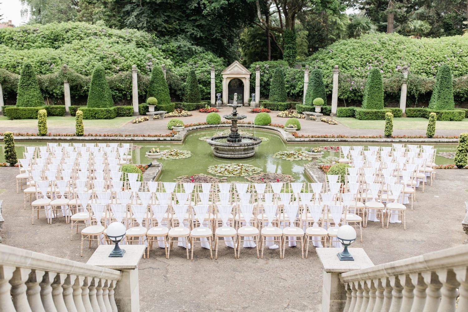 Elegant outdoor wedding ceremony setup with white chairs arranged in curved rows facing a garden fountain