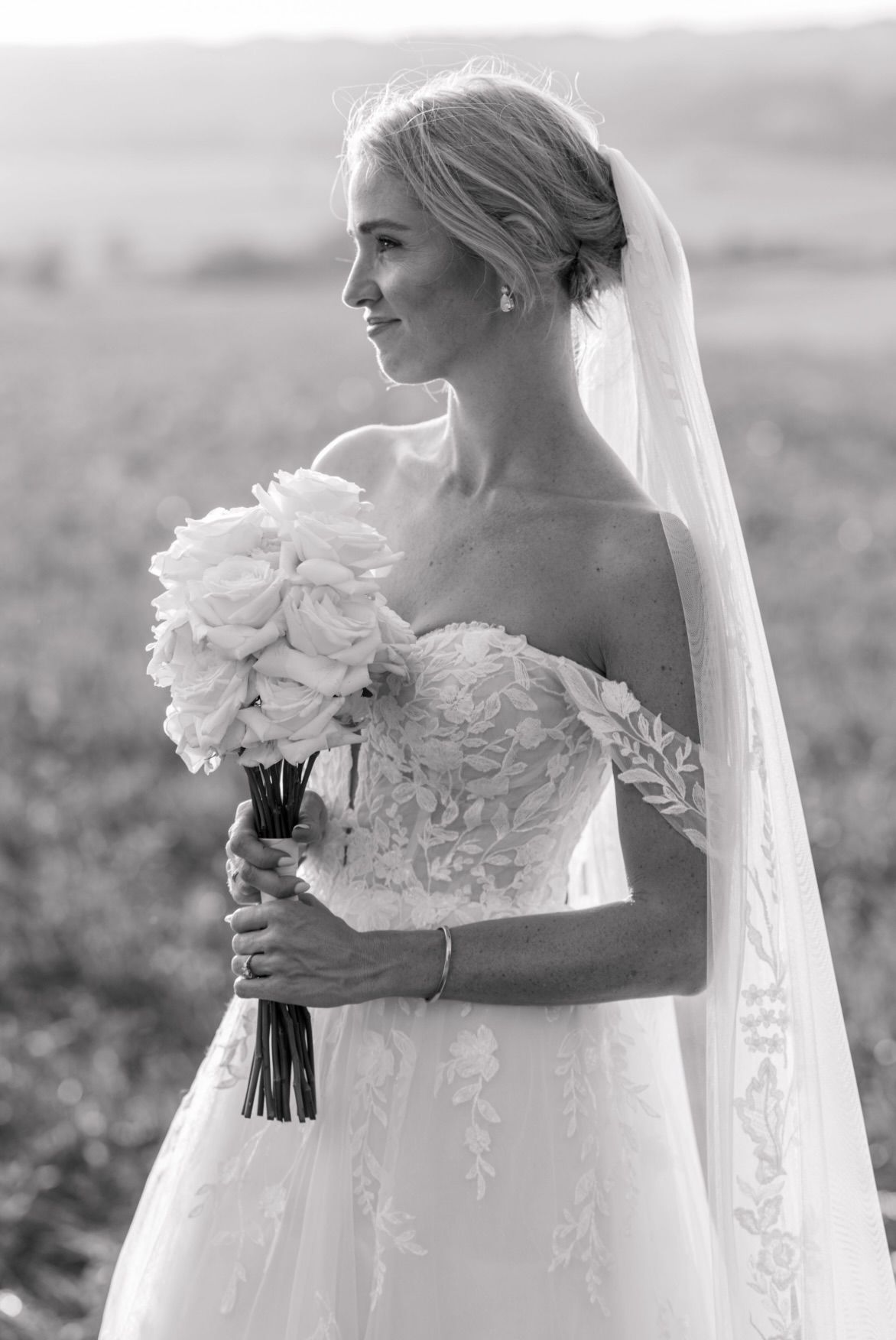 A bride in a strapless lace wedding gown holds a white rose bouquet while posing in black and white photography.