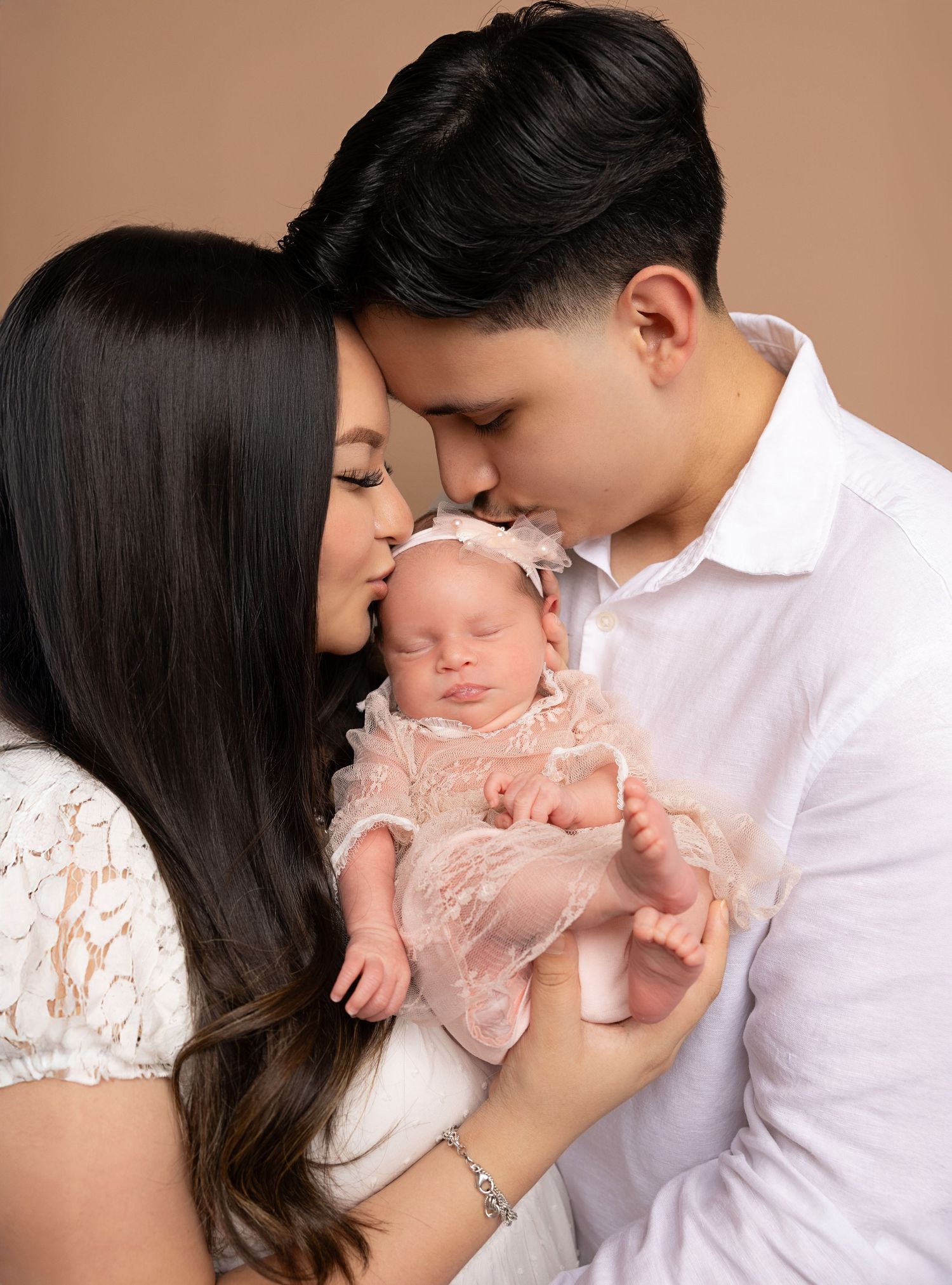 Young parents kissing their sleeping newborn baby girl dressed in a pink lace outfit during a studio portrait session.