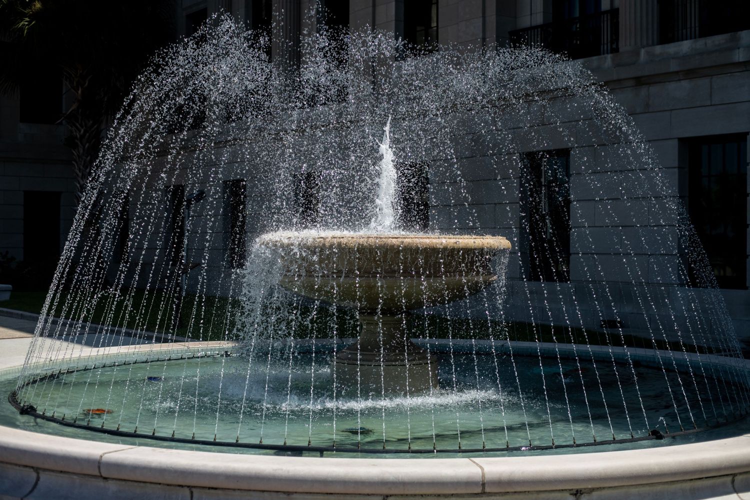 Ornate outdoor water fountain with cascading arcs of water in a circular basin, surrounded by classical architecture.