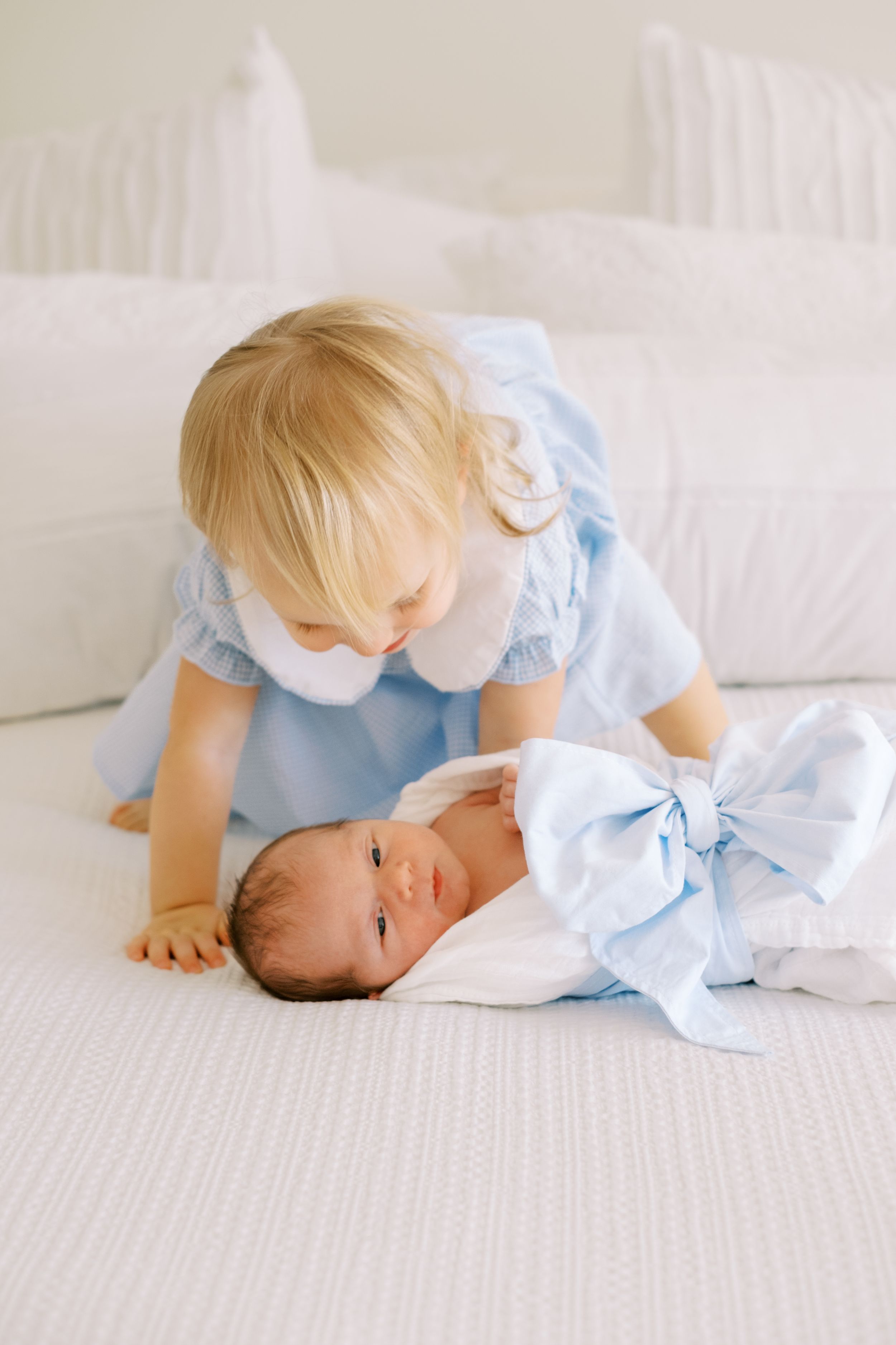 A child in a light blue dress lies on white bedding while looking at something out of frame.