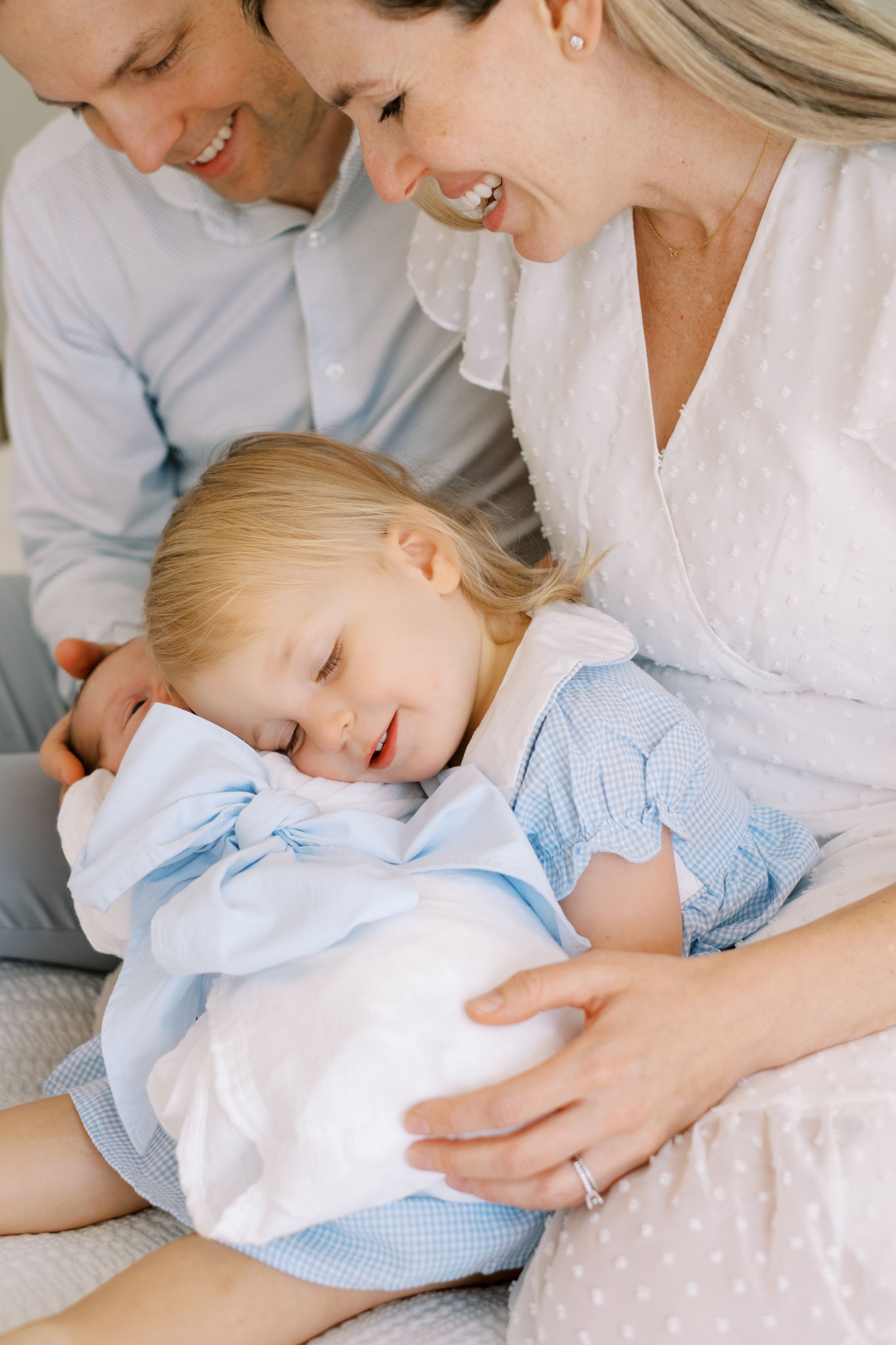 A young child in light blue sleeps peacefully while being held by loving parents in a tender family moment.