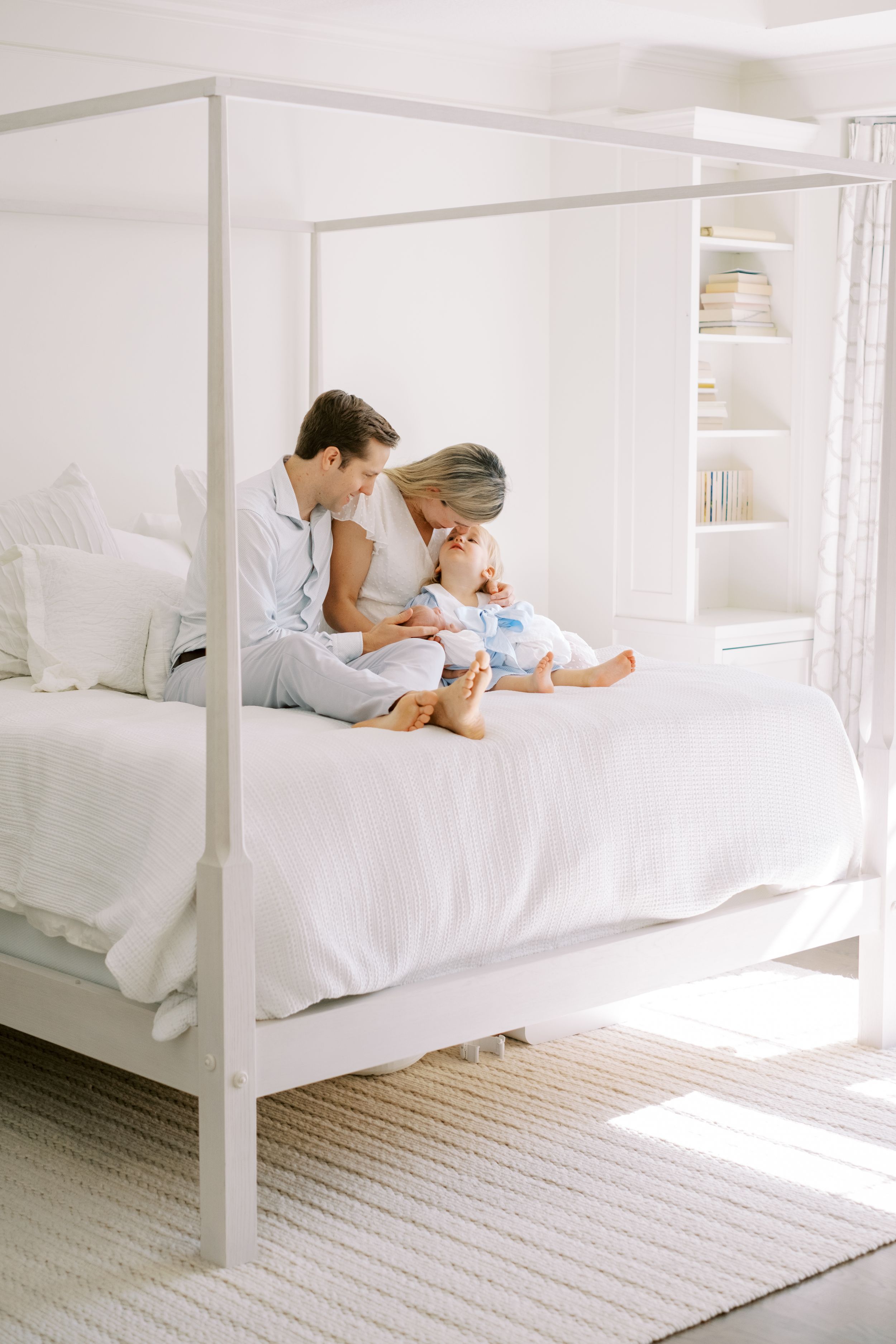 A bright white bedroom with a four-poster bed features people lounging on crisp linens in natural sunlight.