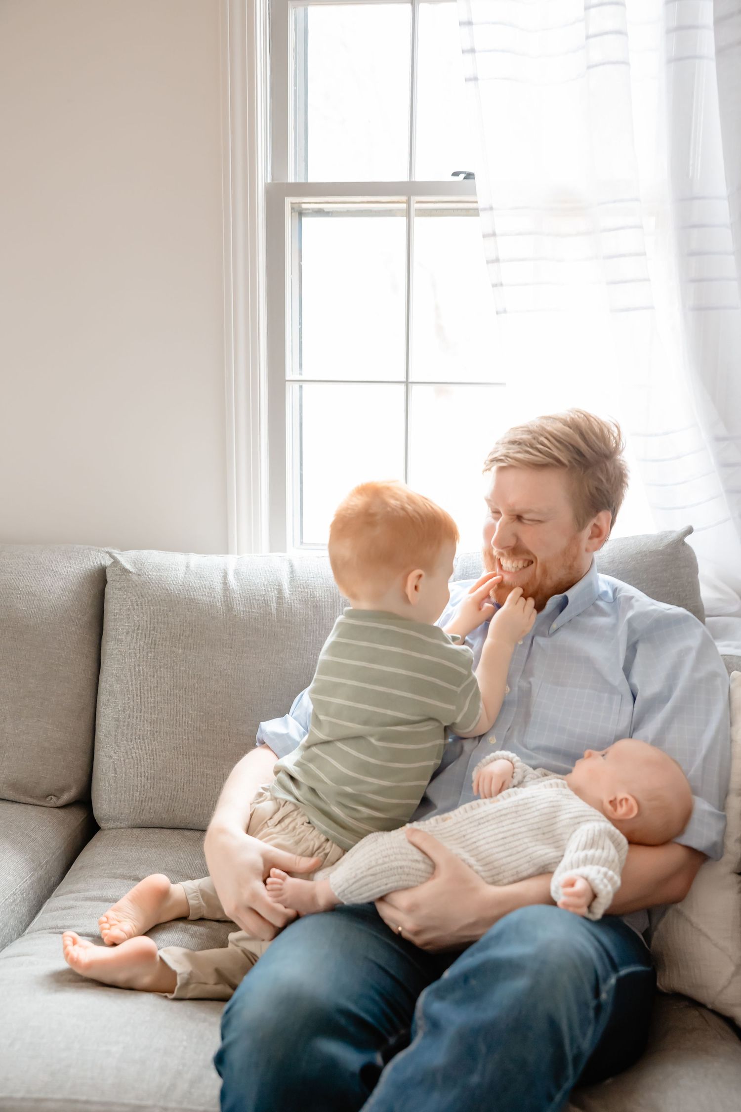 Parent and child share a tender playful moment while relaxing together on a gray couch in a bright room.
