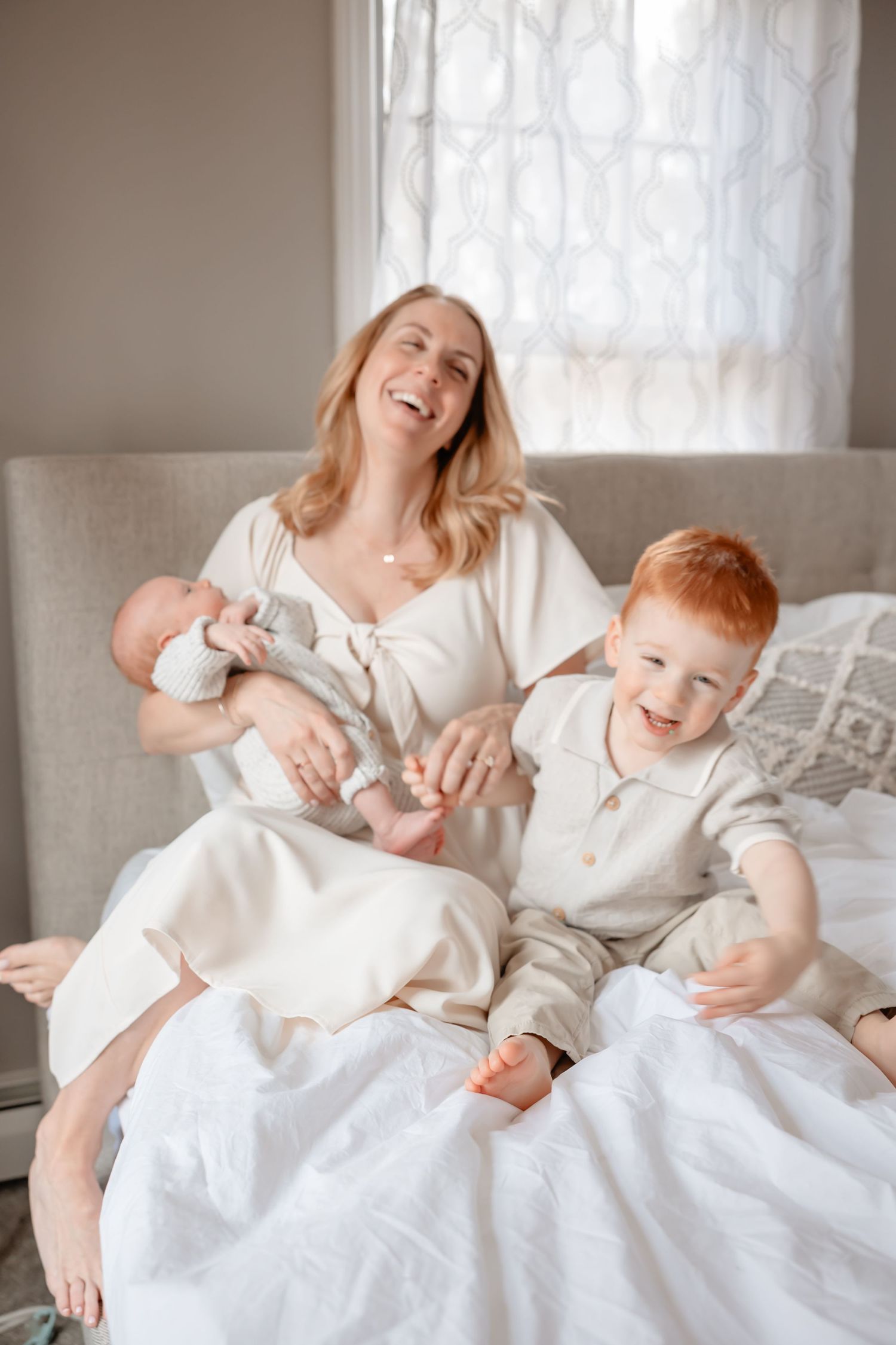 A joyful family moment on a white couch as a mother laughs with her children in matching light-colored outfits.