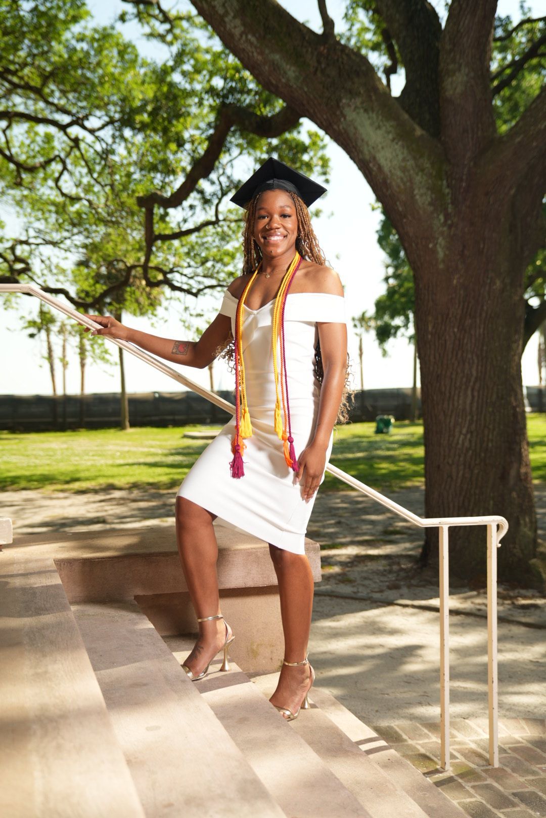 A smiling graduate in a cap, white off-shoulder dress, and colorful honor cords poses on outdoor steps beneath a large oak tree.