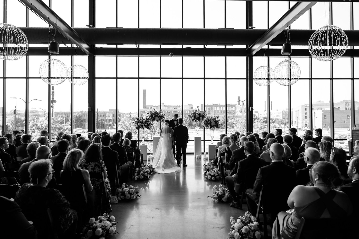 Black and white wedding ceremony photo in elegant venue with large windows, guests seated as couple stands at floral altar.