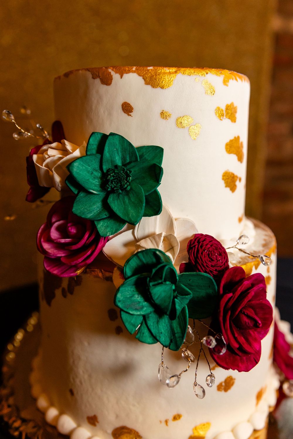 Elegant white wedding cake with gold leaf accents, emerald green and burgundy sugar flowers, and crystal details.