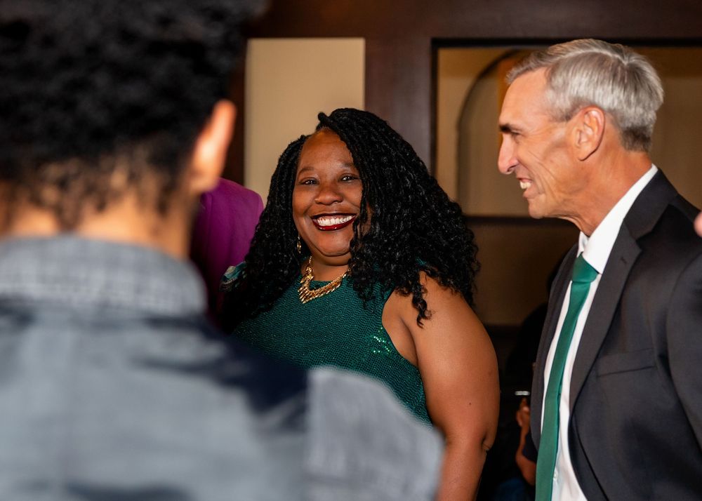 A woman in a green dress laughs joyfully while networking with guests at a formal indoor event.