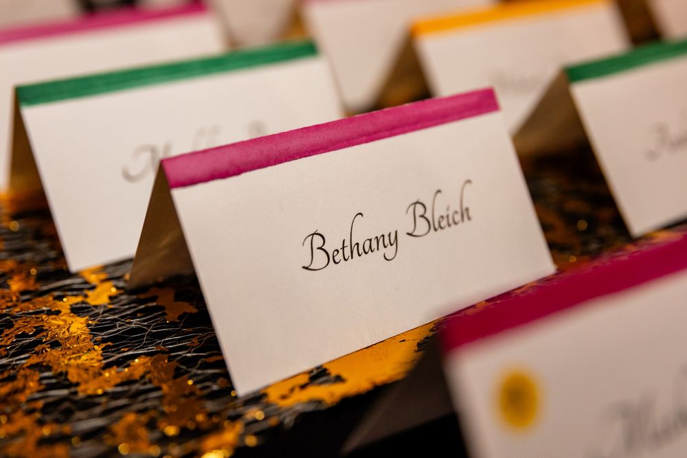 Elegant wedding place cards with colorful borders displaying guest name Bethany Bleich on a glittery gold surface.