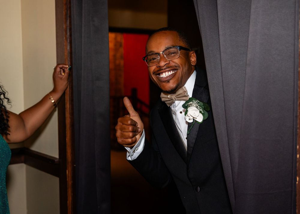 Smiling groom in black tuxedo giving thumbs up while peeking through a curtained doorway at wedding at Blue Sushi in Downtown Omaha, NE