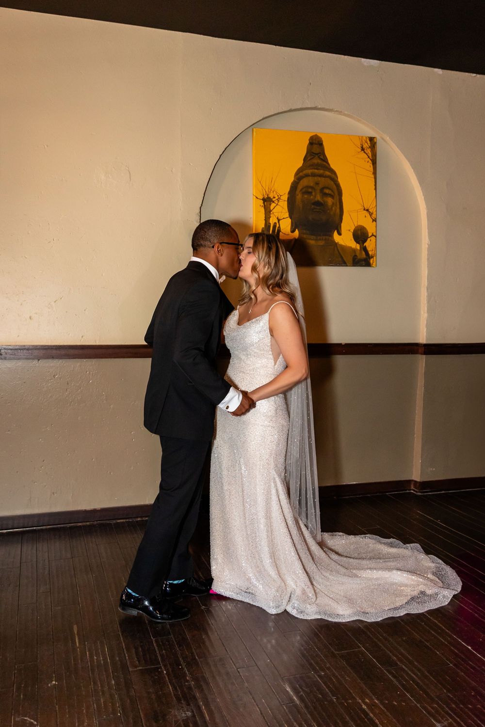 Newlywed interracial couple sharing a kiss in front of a Buddha painting at their wedding reception  at Blue Sushi in Downtown Omaha, NE