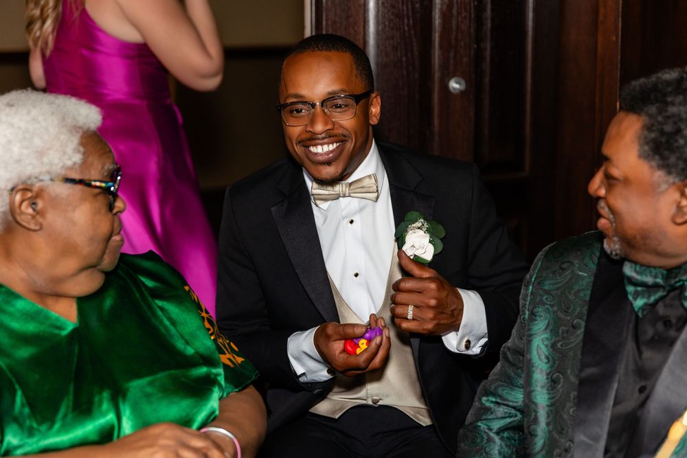 Smiling groom in tuxedo with boutonniere holding a small bowl while seated with wedding guests  at Blue Sushi in Downtown Omaha, NE