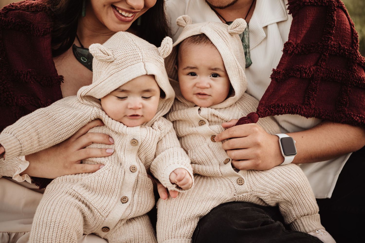 Two babies wearing matching cream knit outfits and bonnets sit on laps in a family portrait.
