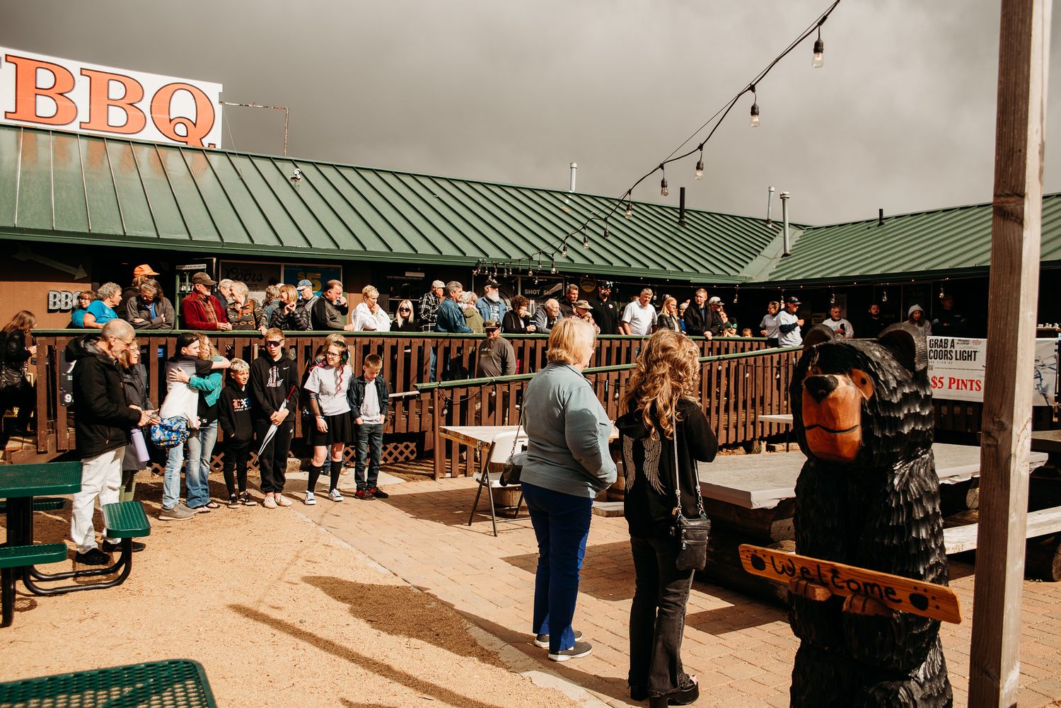 Crowd gathered at an outdoor BBQ restaurant patio with string lights on a cloudy day.