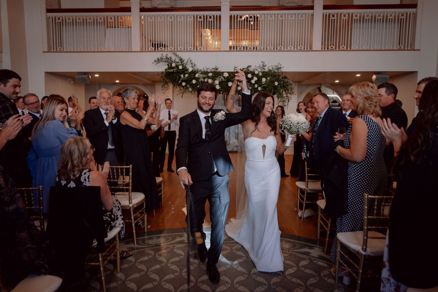 Newlyweds walking down the aisle while guests celebrate in an elegant ballroom venue.