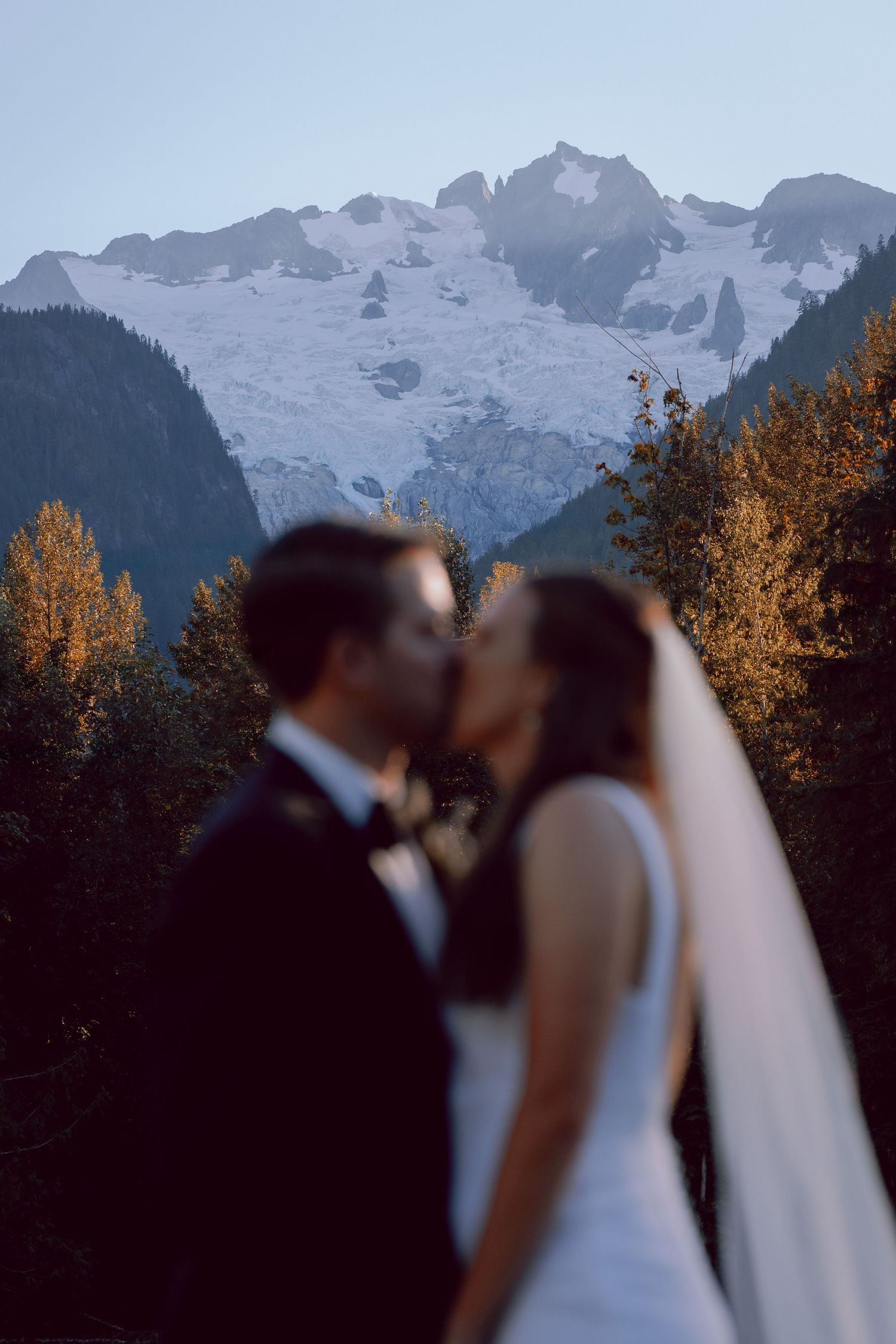 A couple shares a romantic moment in front of snowy mountain peaks and fall foliage at sunset.