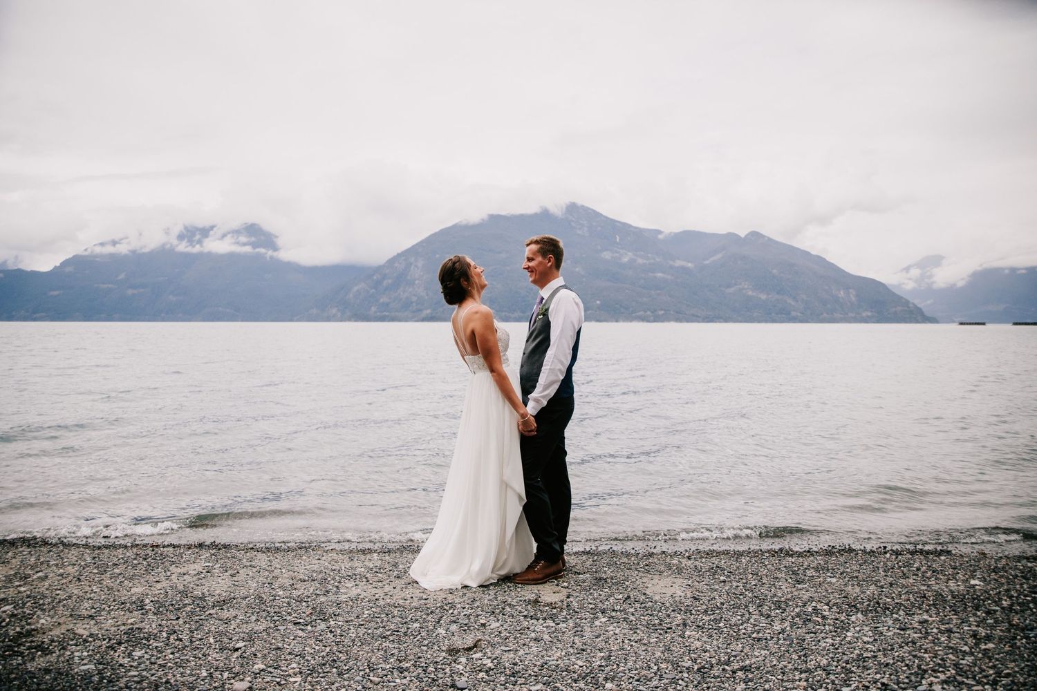 Bride and groom standing on beach with misty mountains in background.