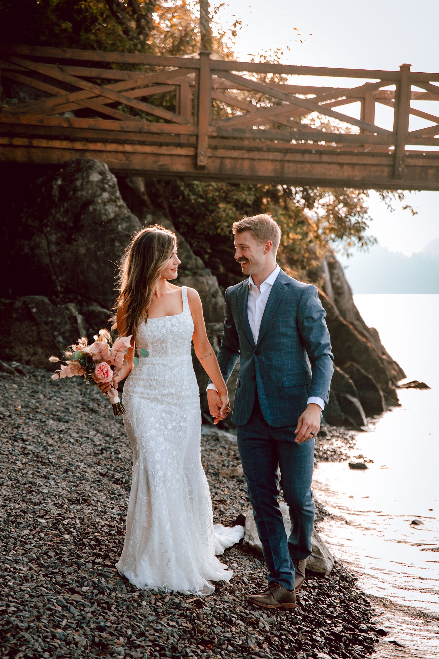Newlyweds walk hand in hand along rocky beach shore with wooden bridge in background.