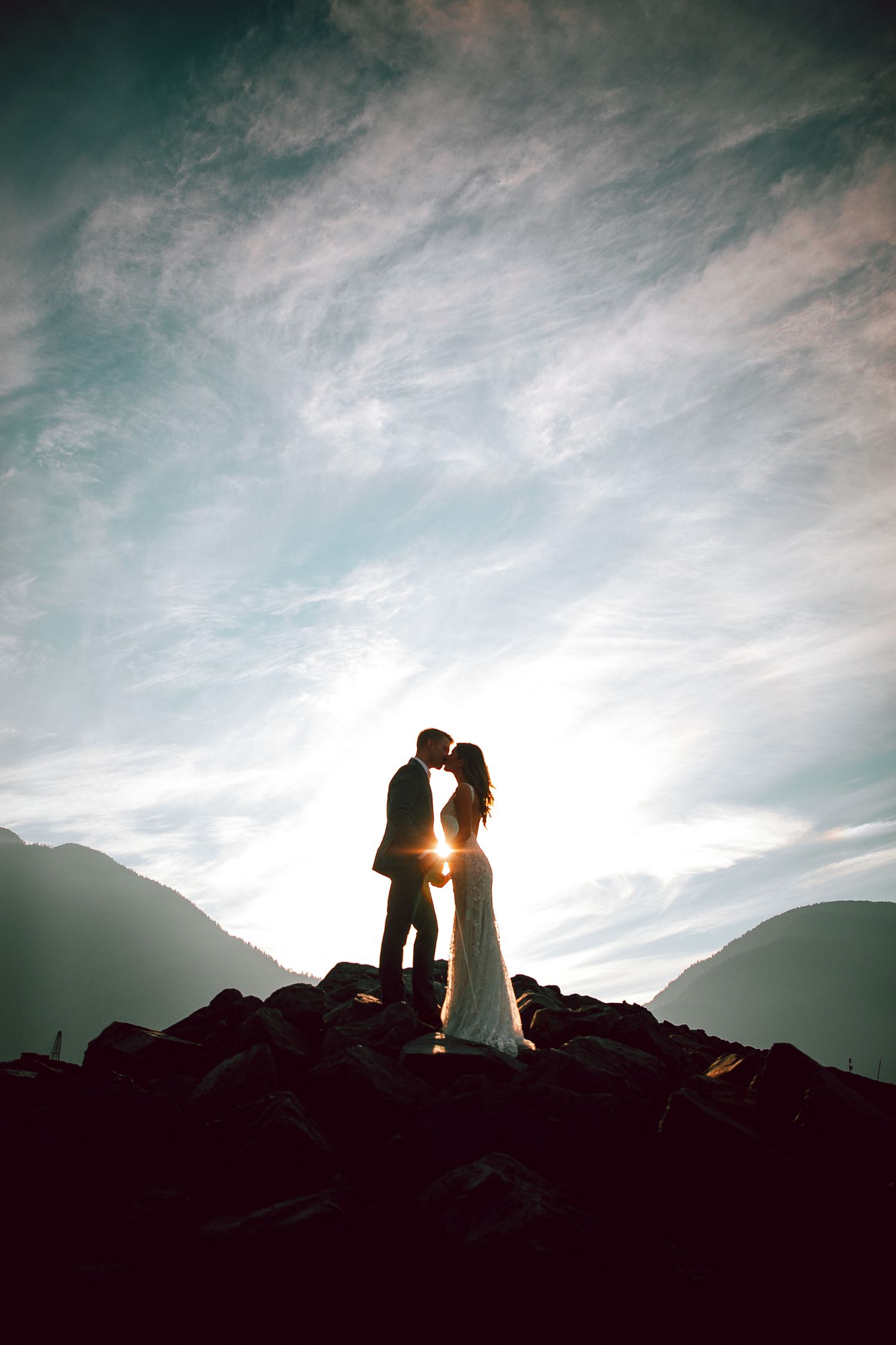 Silhouette of newlyweds standing on rocky outcrop against dramatic sunset sky with mountains.